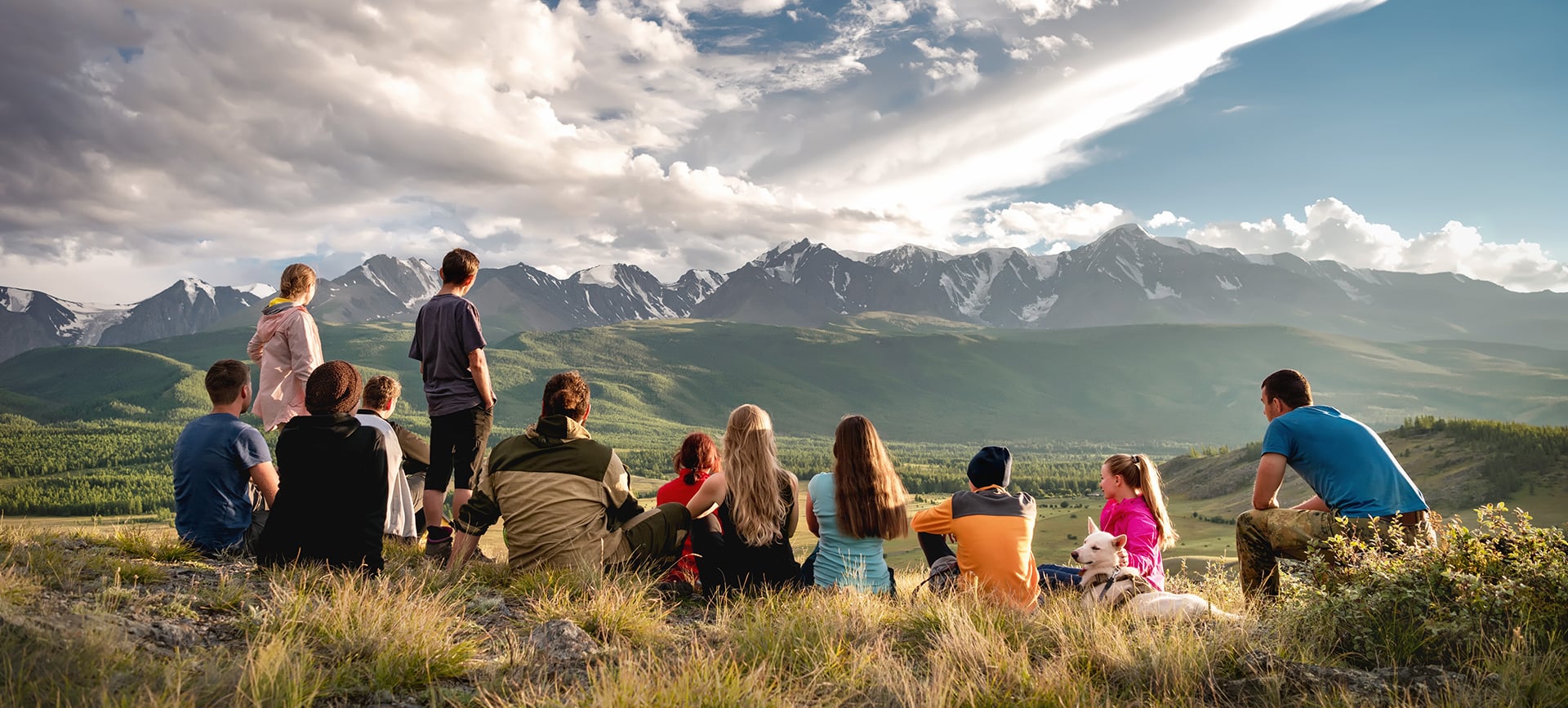 Gruppo di viaggiatori seduto su una cresta erbosa davanti a una catena di montagne innevate